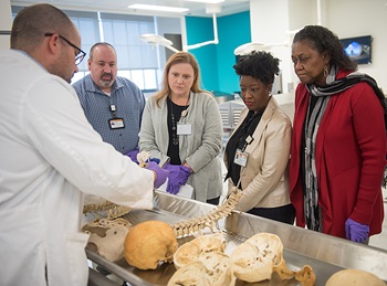 A group of inpatient medical coders surround a table with a skeleton on it and listen as man in white coat explains the bone he's holding