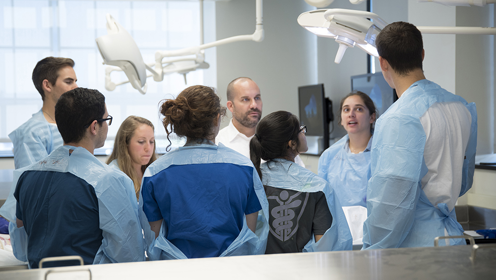 First-year medical students wear blue paper gowns and stand around a table during anatomy lab