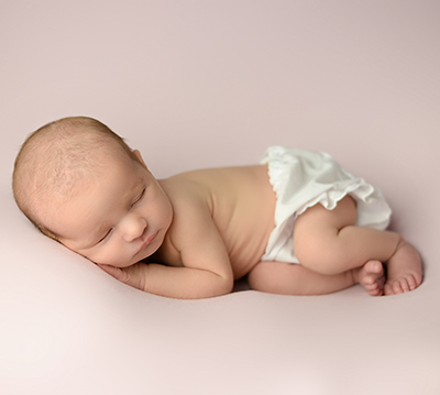 Newborn baby wearing frilly white cloth diaper sleeps against a soft pink background with its hands tucked under its head and its knees drawn up 