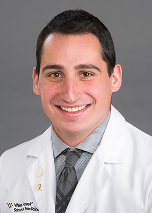 A young man with dark hair and dark eyes, wearing a white coat, smiles at the camera