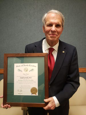 An older man in a red tie and dark suit smiles as he holds a framed plaque