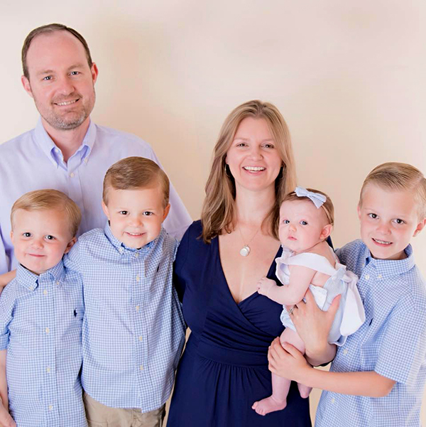 Family photo of mother, father, three boys and a baby. Father and boys are in light blue shirts, mom in a navy dress and baby in white