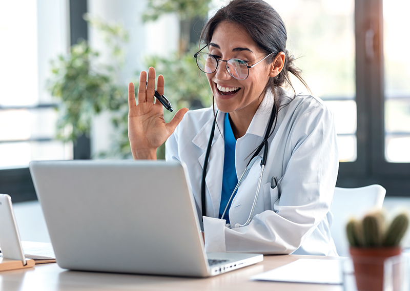 Woman in a white coat and stethoscope with dark hair pulled back smiles and waves at laptop screen in well-lit office