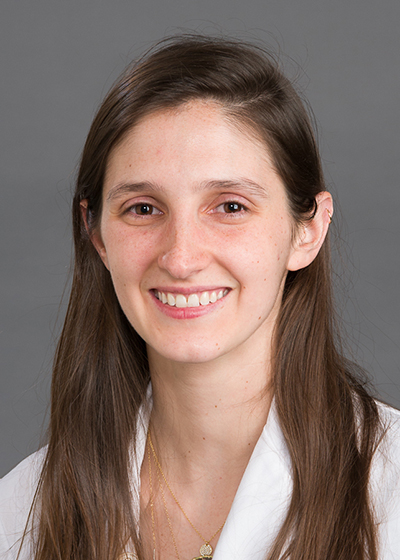 Young woman with long dark hair and brown eyes, wearing a white coat, smiles at camera