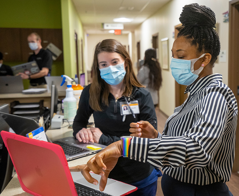 Two women wearing face masks stand in the hallway of a health clinic and look at a laptop screen