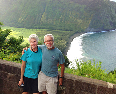 A man and woman, both white-haired and wearing shorts and T-shirts, stand in front of a rock wall that overlooks a black sand beach and vegetation-covered mountain