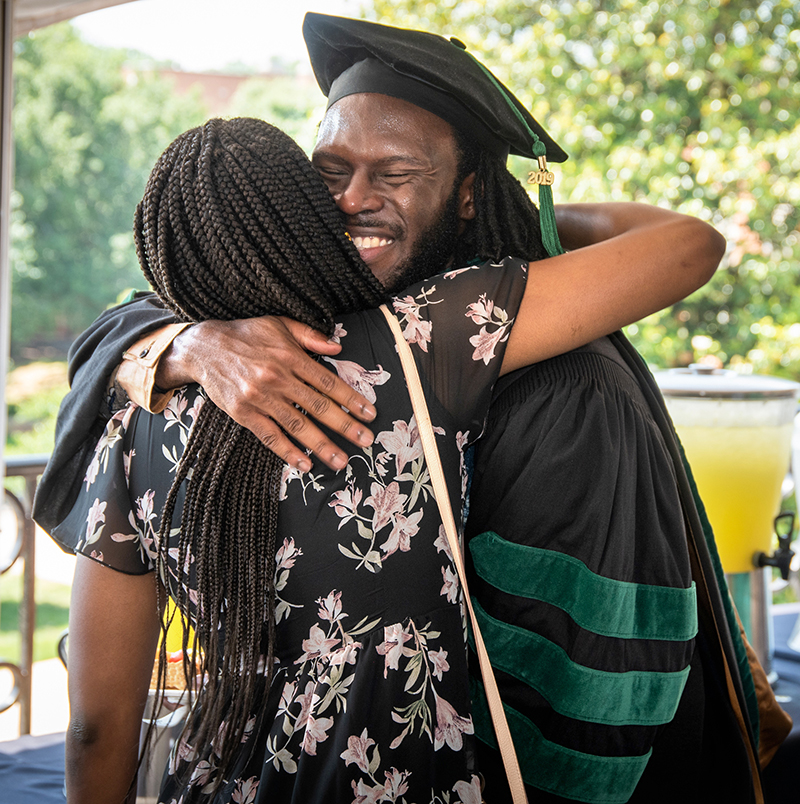 An African-American man wearing black graduation robes with green velvet stripes on the sleeves hugs a woman outside