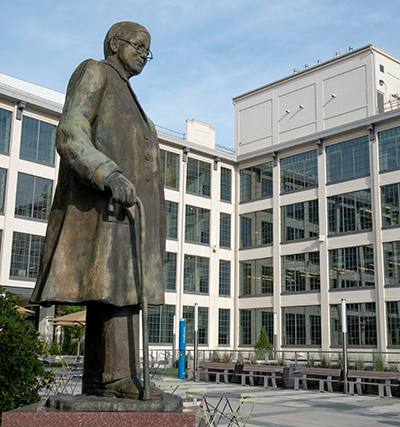 Courtyard of concrete building with large windows against a blue sky, with a statue of a man in the foreground 