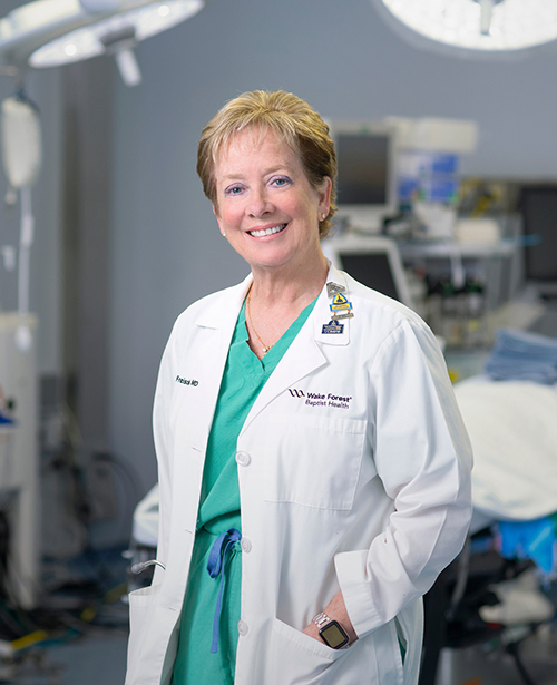 A woman with short blondish hair wears green OR scrubs and a white coat and stands in an operating room