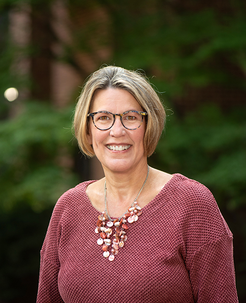 A woman with chin-length hair, glasses and a burgundy sweater stands outdoors and smiles at the camera