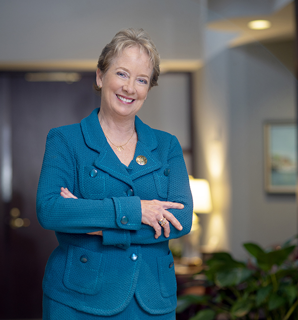A woman with short blond hair wearing a teal suit smiles as she stands in an office