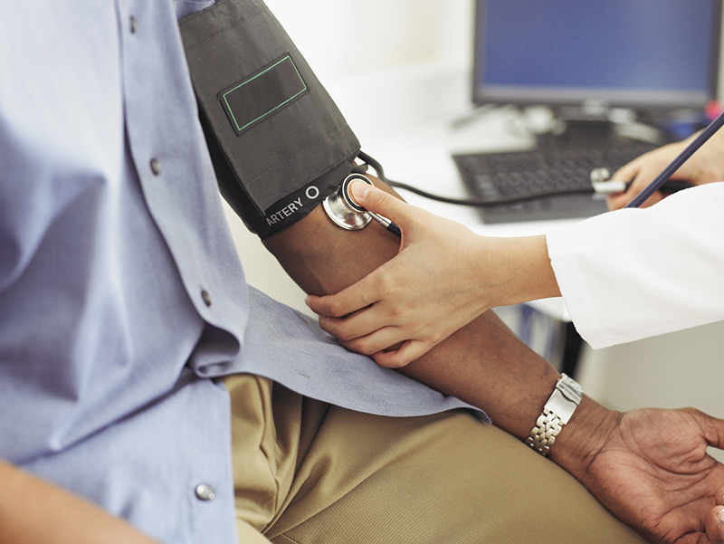 A seated man in blue shirt and tan pants gets his blood pressure taken by person wearing a white coat