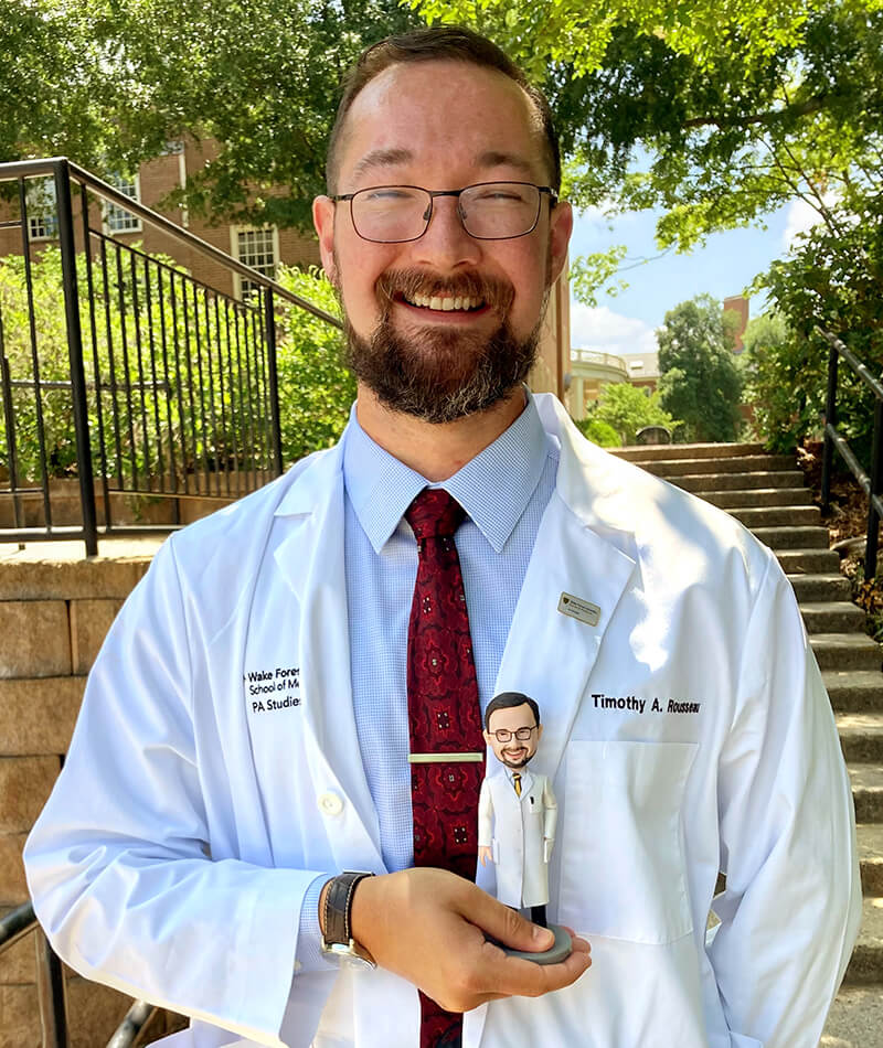 A young man wearing a lab coat holding a bobblehead doll and smiling at the camera.