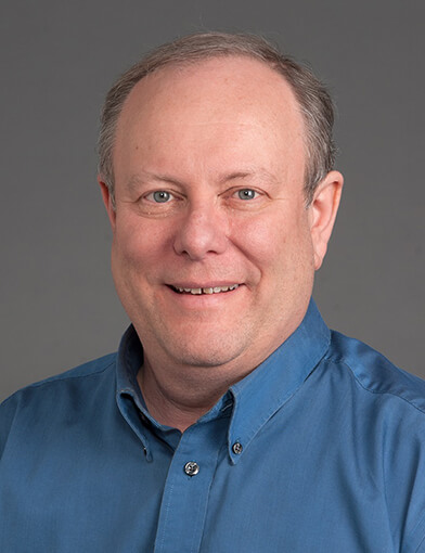 An older man wearing a blue shit looking at the camera.