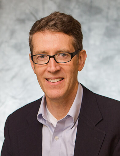 A man wearing glasses and a suit jacket smiling at the camera.