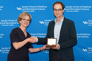 Two people standing in front of a blue background and shaking hands over an award.
