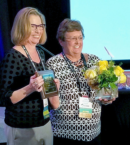 Two women, one holding an arrangement of yellow flowers and the other holding a plaque, stand in front of a screen