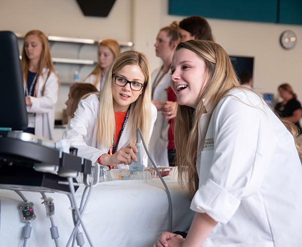 Two young Caucasian women wearing white lab coats talk as they look at a ultrasound monitor