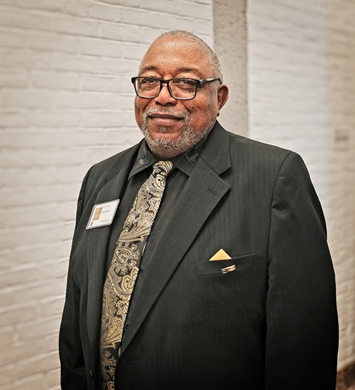 An older African-American man in a black suit and wearing glasses smiles at the camera