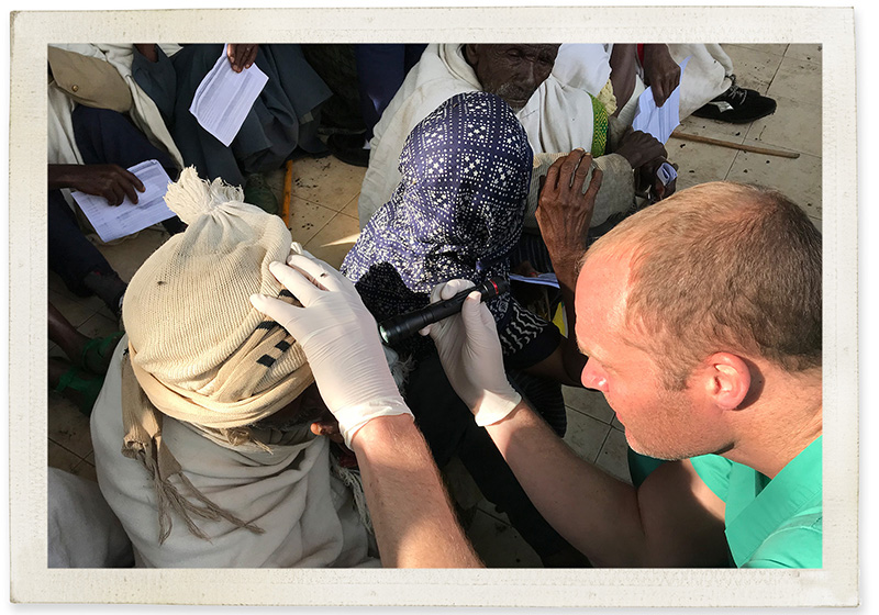 Caucasian man in scrubs uses a light to examine the eyes of a of a turbaned man