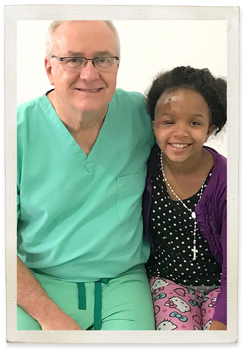 Older Caucasian man in green scrubs sits with a dark-skinned young girl and both smile at the camera