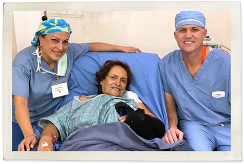 A man and woman wearing blue scrubs sit beside an older female patient on a hospital bed