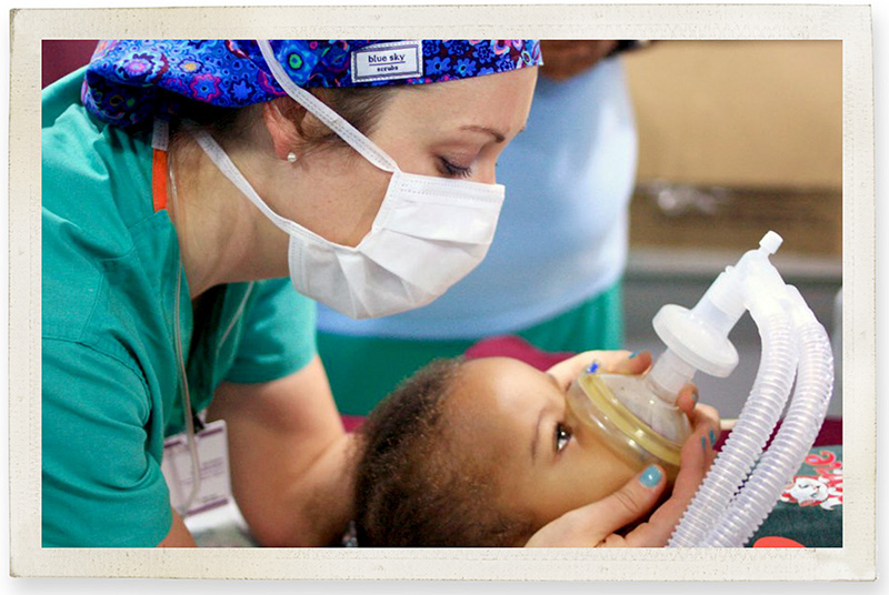 Woman with brown hair, a blue hair mask and a surgical mask, wearing green scrubs, leans over a young child wearing a breathing mask