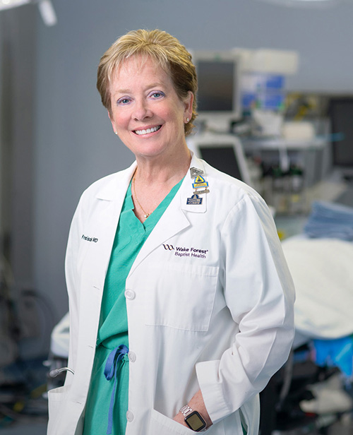 Caucasian woman with strawberry-blond hair wearing green scrubs and a white coat stands in an operating room