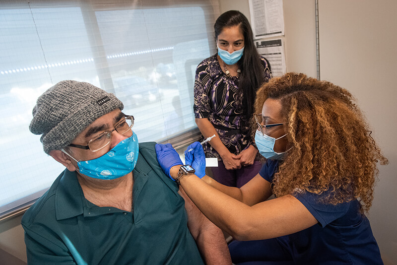 A medical professional wearing a mask giving a shot to an older man wearing a mask while another woman watches.