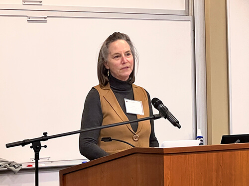 A woman wearing a long-sleeved shirt and brown vest speaking at a podium.
