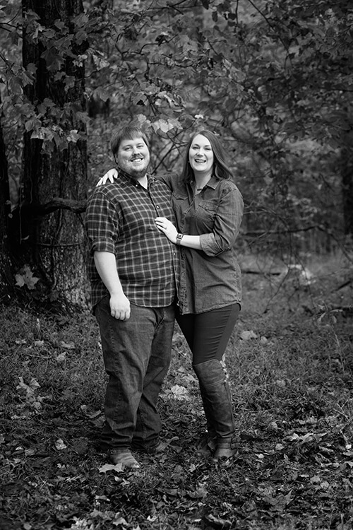 A man and woman standing in the woods smiling at the camera.