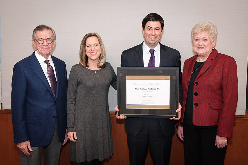 Two women and one man standing around another man holding an award.
