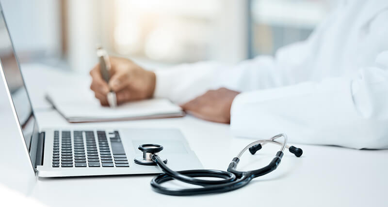 A medical professional writing notes beside a laptop and a stethescope.