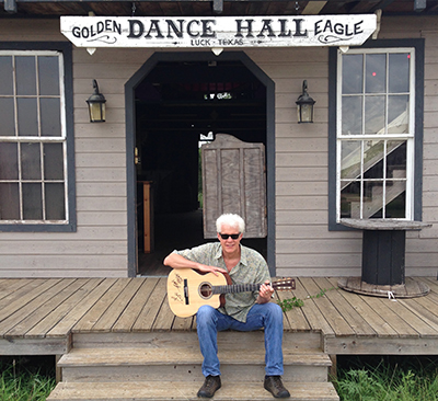 A white-haired man sits on wooden steps with a guitar in his hands