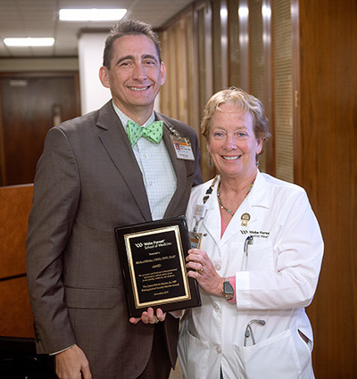 A man in a bow tie smiles as he receives a plaque from a woman in a white coat