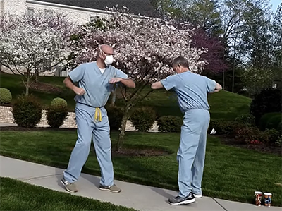 Two men in light blue scrubs and masks touch elbows on a sidewalk in front of a house