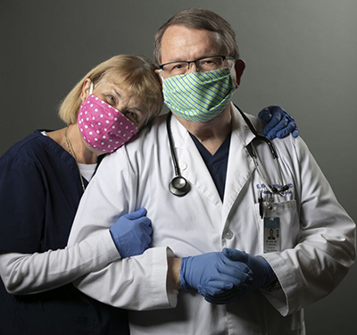 A woman in navy scrubs, mask and gloves, puts her head on the shoulder of a masked, gloved man in a white coat
