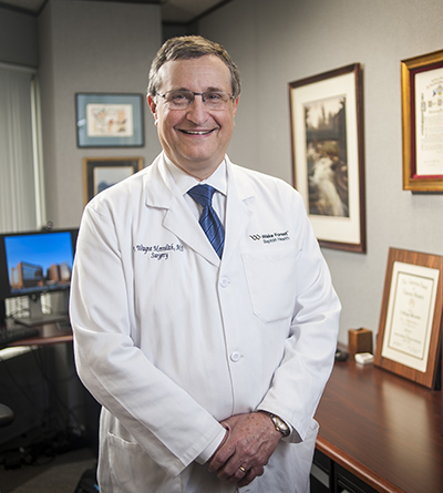 An older Caucasian male wearing a white coat, tie and glasses stands in an office