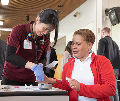 Seated African American woman in red cardigan has finger pricked by Asian American female medical student