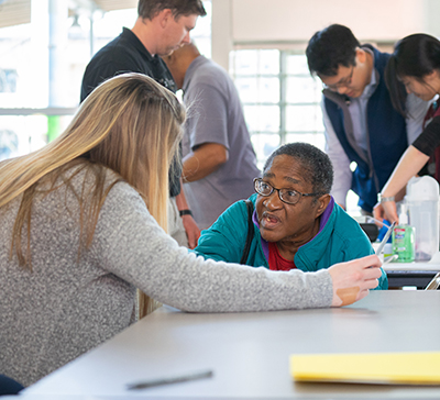 Seated older African American woman talks to medical student with long blonde hair