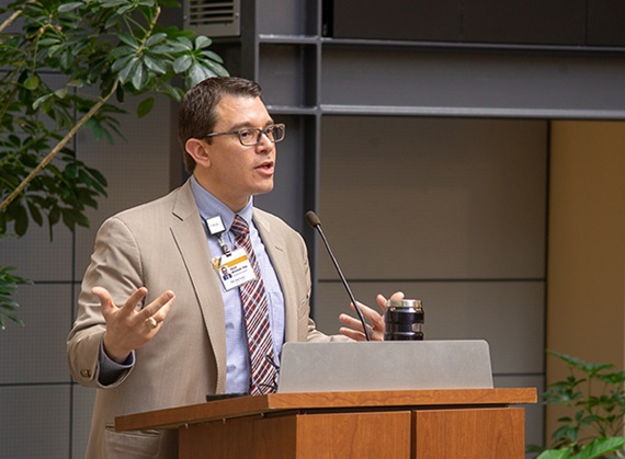 Caucasian male in jacket and wearing glasses speaks at a podium