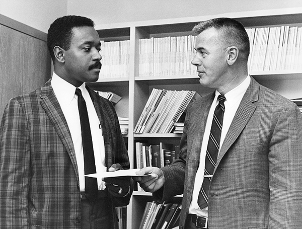 Two men, one Black and one white, stand in front of bookshelves in a black-and-white photo, circa 1970s