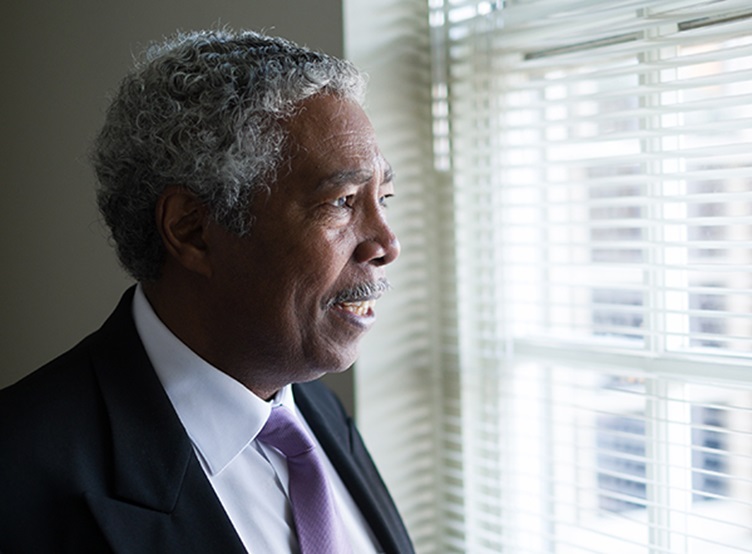 An older Black man stands at a window, looking outside