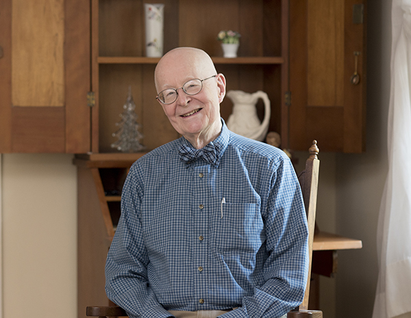 A bald, older Caucasian man wearing glasses and a bow tie smiles at the camera