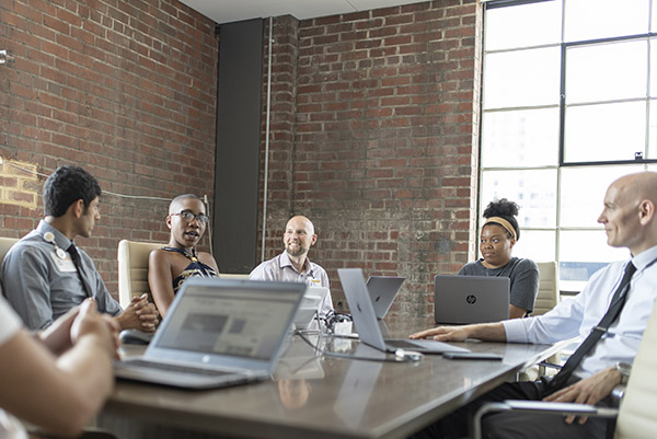 A diverse group of people sit around a conference table with laptops in a room with exposed brick and large windows