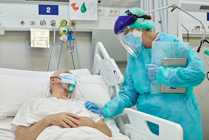  Female medical professional wearing protective garment, face shield, surgical mask, and gloves rests her hand on shoulder of older male patient in a hospital bed 