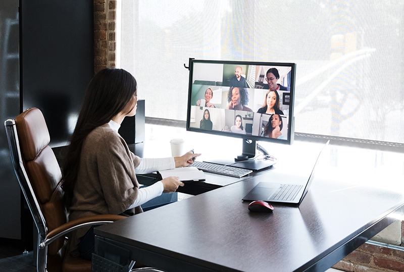 Woman with long dark hair sits at office desk on a video conference call in front of large window