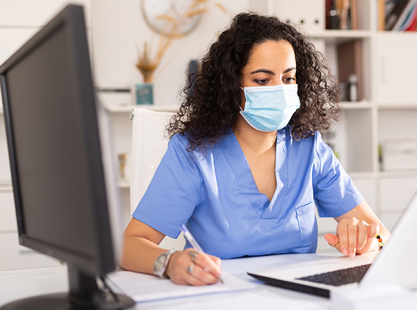 A young black woman in blue scrubs and wearing a disposable paper mask and looks down at a tablet