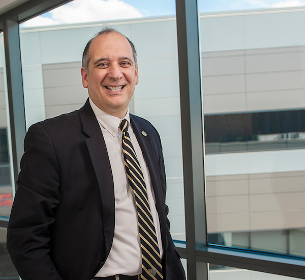 Middle-age white man in jacket and tie stands in front of large window and smiles