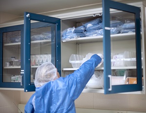 Person in blue protective gown and hair net reaches up to remove supplies from blue cabinets with glass doors